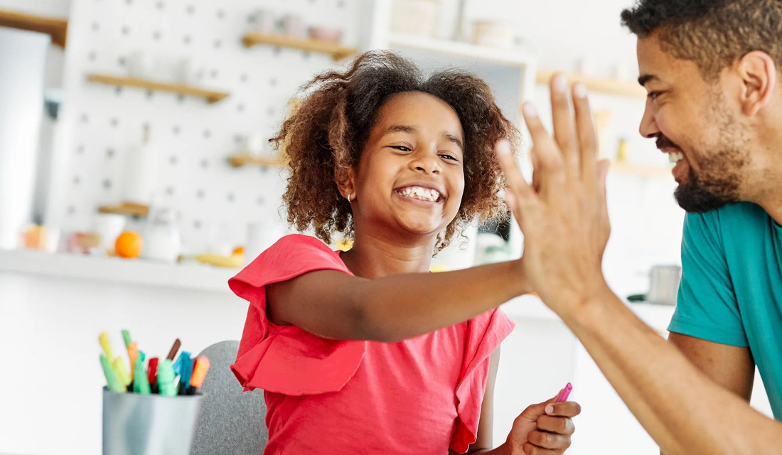 A father and daughter sitting at a table, smiling and giving each other high fives while doing schoolwork together.