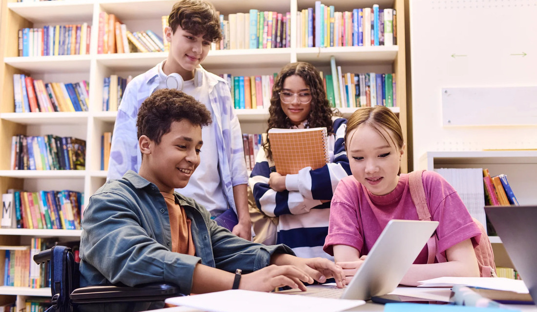 A group of students gathered in a library, focused on a laptop screen while discussing their work.