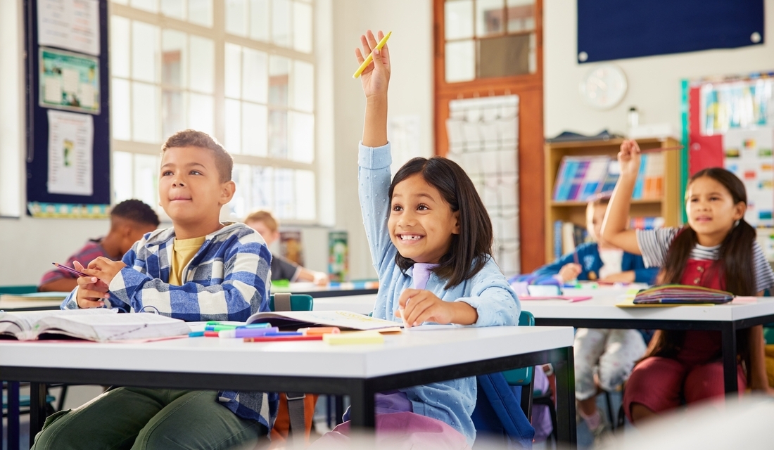 Students in a classroom, sitting at desks with hands raised, indicating their desire to contribute to the discussion.