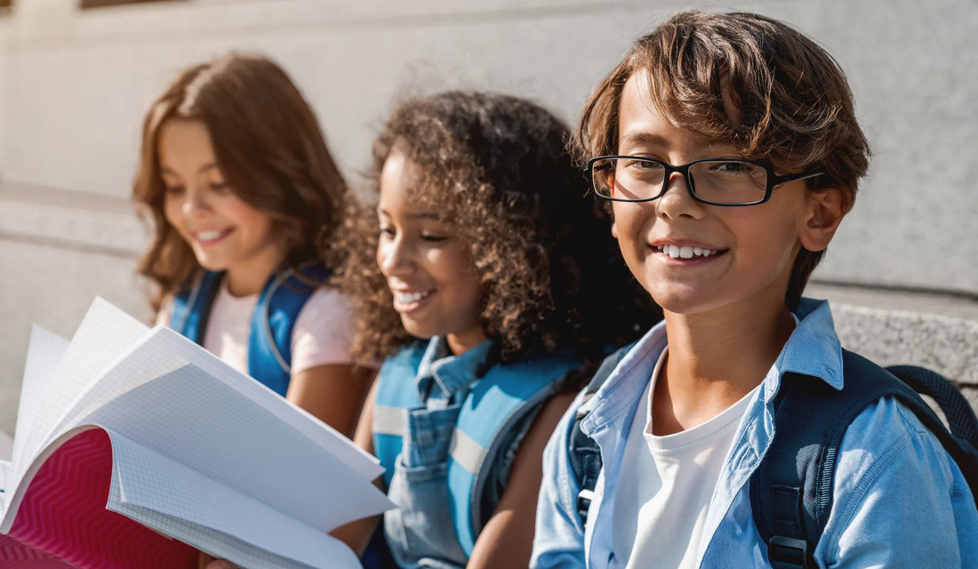 Three bespectacled children are seated on a bench, displaying cheerful expressions as they interact with one another.