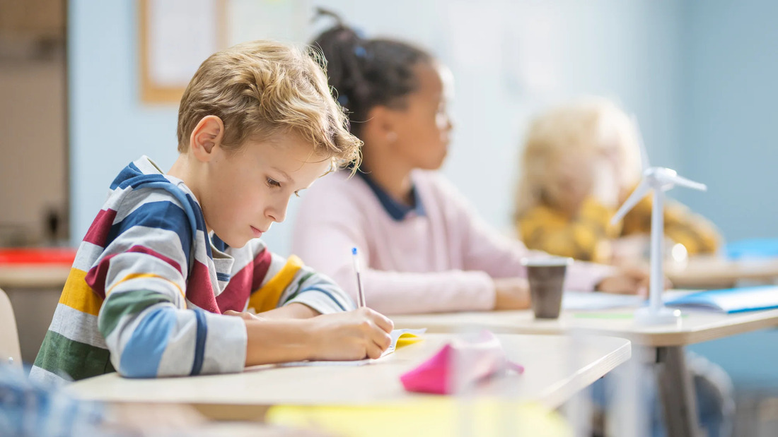 A young boy sits at a desk in a classroom, focused on writing in a notebook.