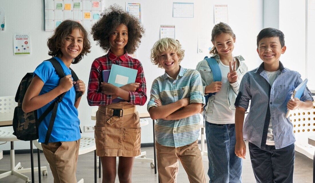 A group of children stands in a classroom, each holding books and engaged in a learning activity.