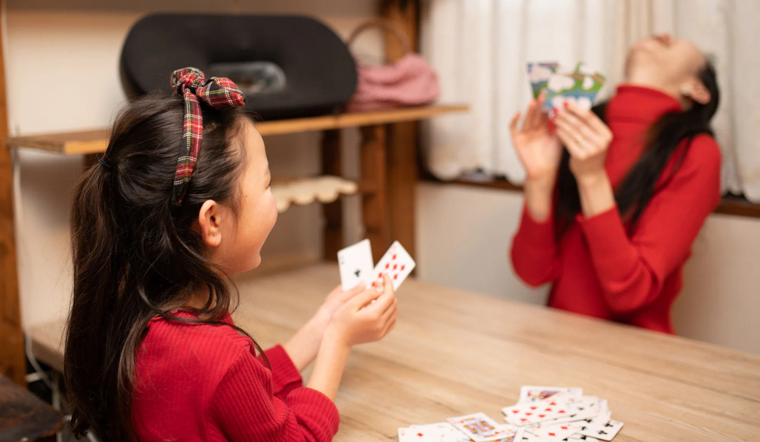 A young girl and her mother sit at a table, smiling and playing cards together in a cozy indoor setting.