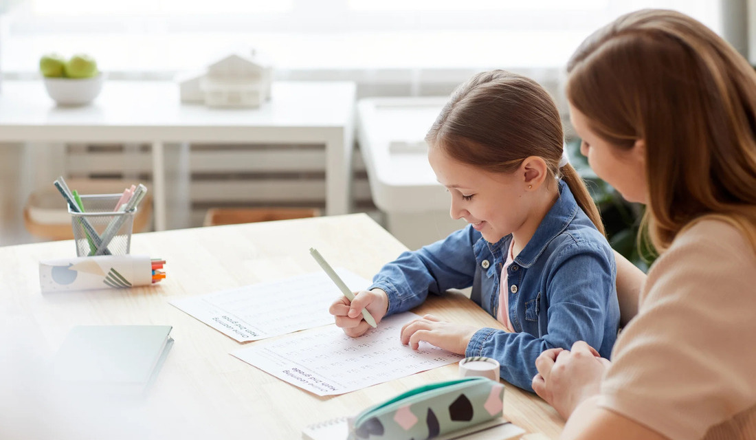 A woman and a little girl are seated at a table, engaged in writing activities with notebooks and writing tools.