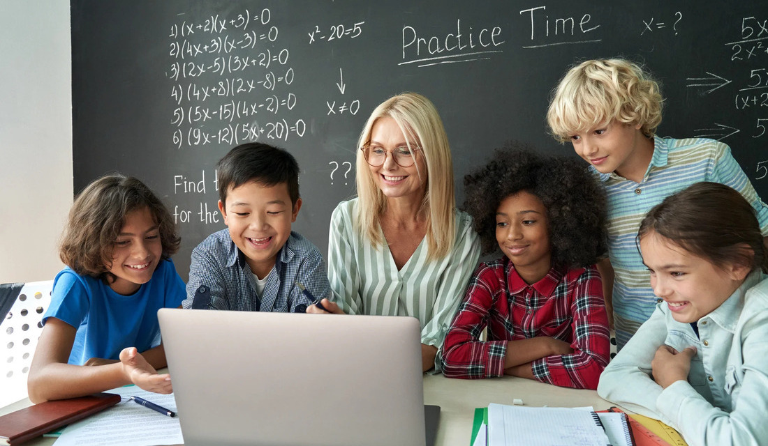 A group of children and their teacher observe a laptop screen, participating in a collaborative educational session.
