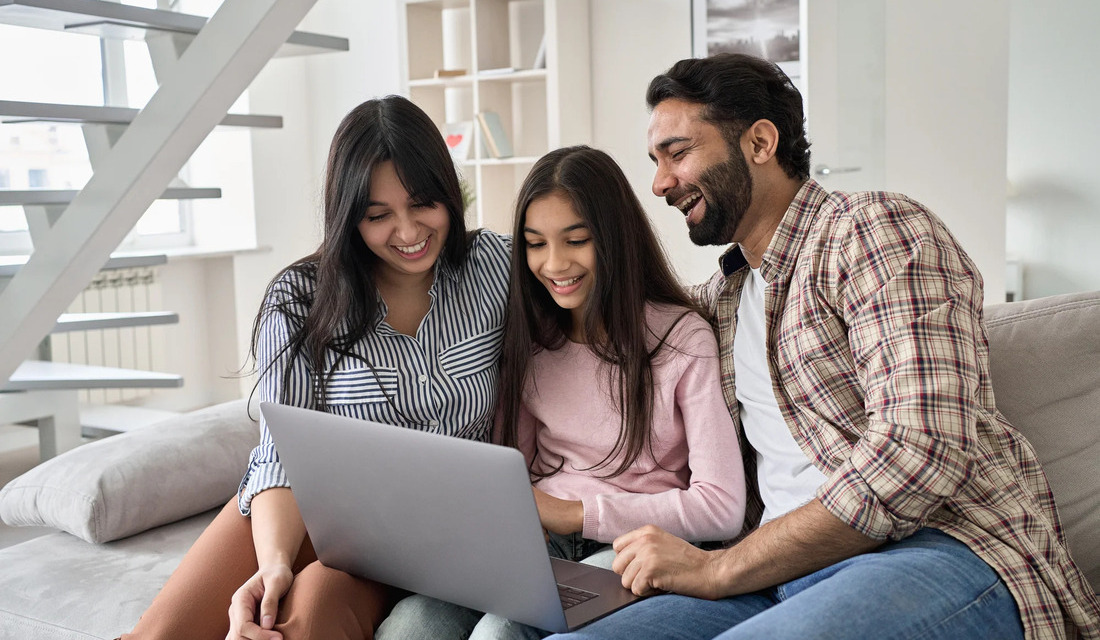 A family of three relaxes on a couch, focused on a laptop, creating a warm and interactive atmosphere.