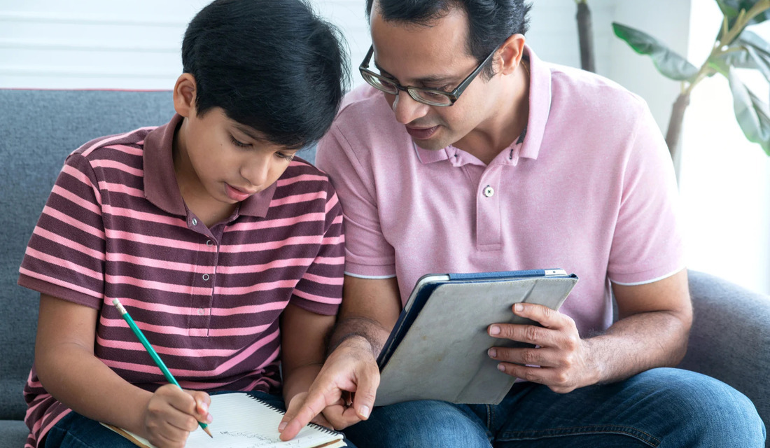 A father assists his son with homework, both engaged and seated comfortably on a couch at home.