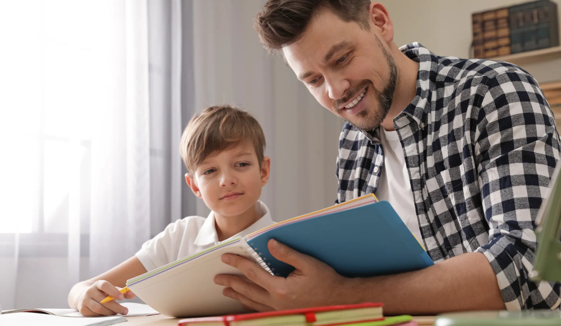 A man and a boy are at a table, both looking at a book, highlighting a bonding moment through reading.