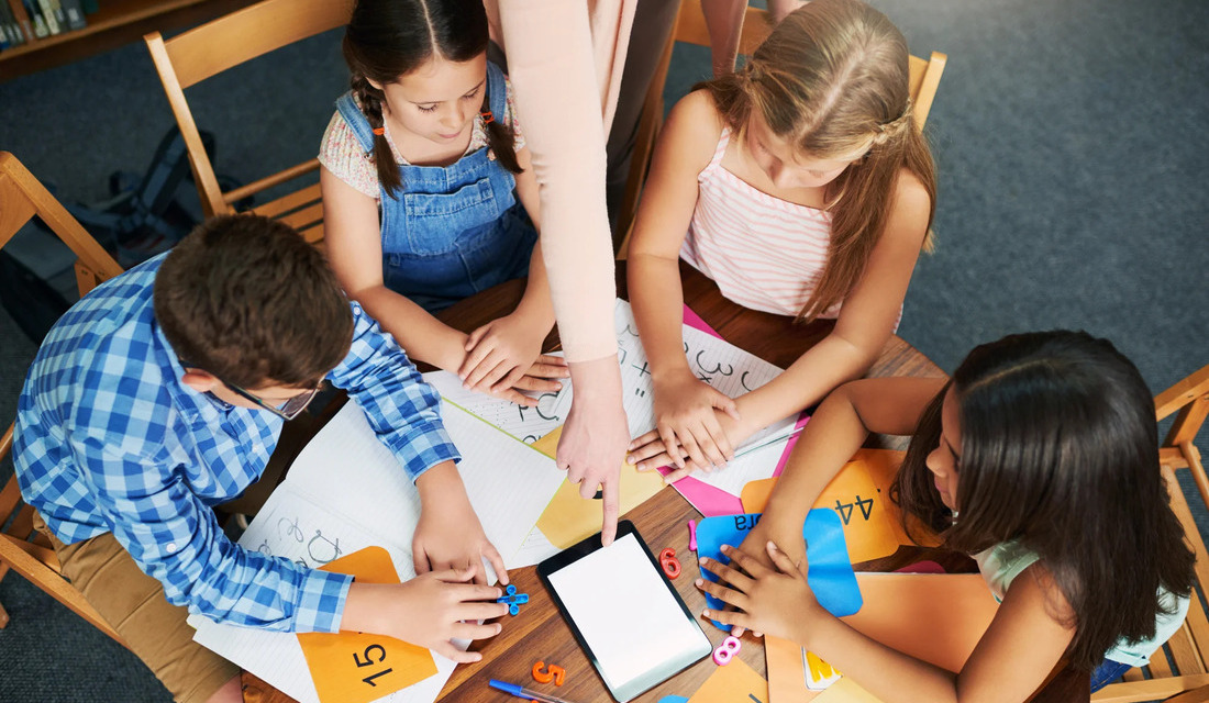 Children gathered around a table, utilizing a tablet and paper for educational tasks and interactive play.