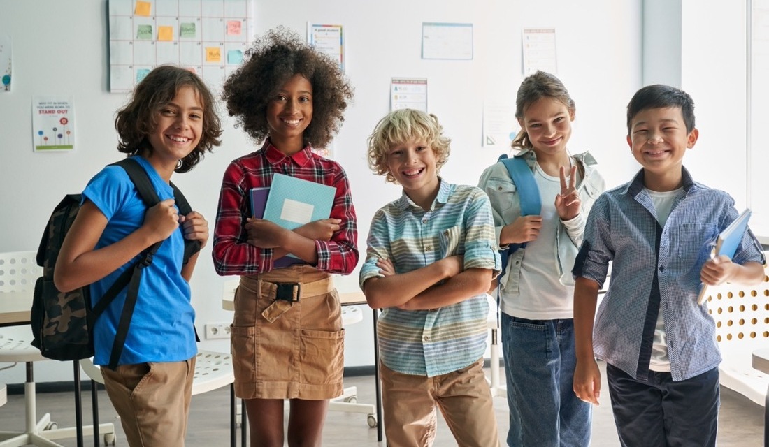 A group of children in a classroom, each wearing backpacks and engaged in conversation.