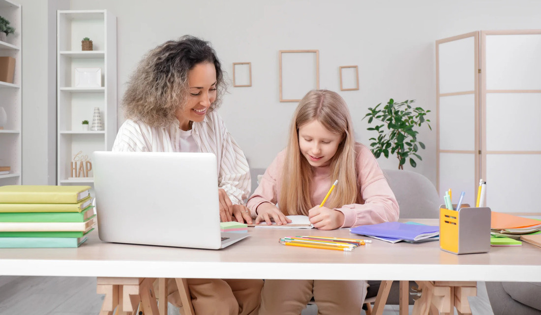A woman and a girl are seated at a table, engaged in an activity using a laptop.