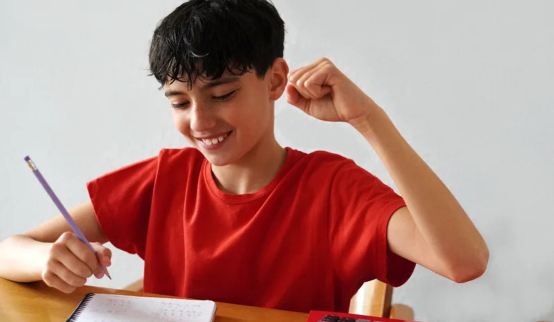 A boy in a red shirt sits at a desk, raising his fist while focused on his homework.