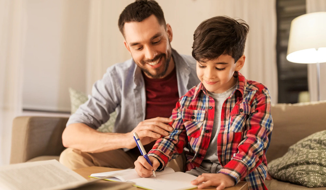 A man and a young boy are seated on a couch, engaged in writing activities with notebooks.