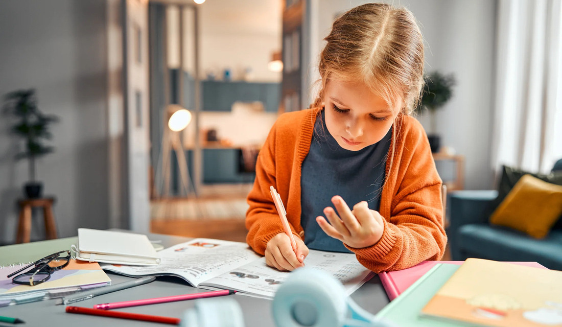 A young girl sits at a table, focused on writing in her notebook with a pencil in hand.