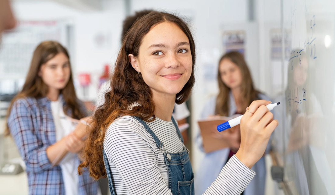 A girl writes on a whiteboard while other students observe and engage in a classroom setting.