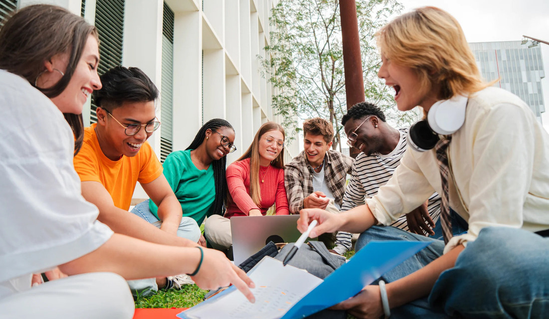 A group of students sitting on grass, collaborating on a laptop in a sunny outdoor setting.
