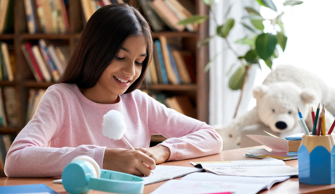 A girl sits at a desk, smiling at a white teddy bear placed beside her, creating a cozy and playful atmosphere.