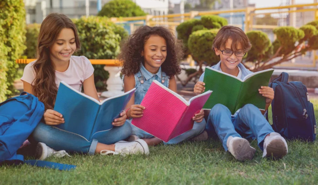 Three children sitting on green grass, each engrossed in reading their own book under a clear blue sky.