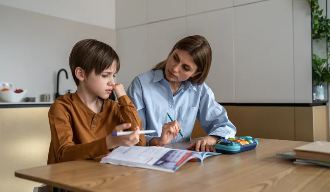 A woman and a boy sit at a table, engaged in writing or drawing in a notebook together.
