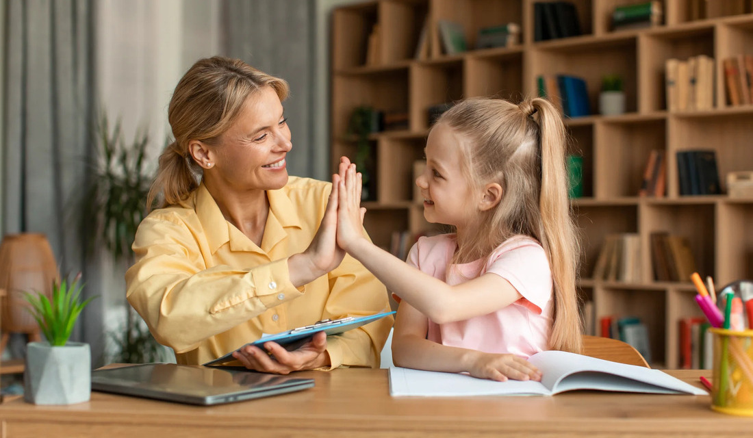 A woman and a little girl at a desk, both with their hands raised, expressing excitement or eagerness.