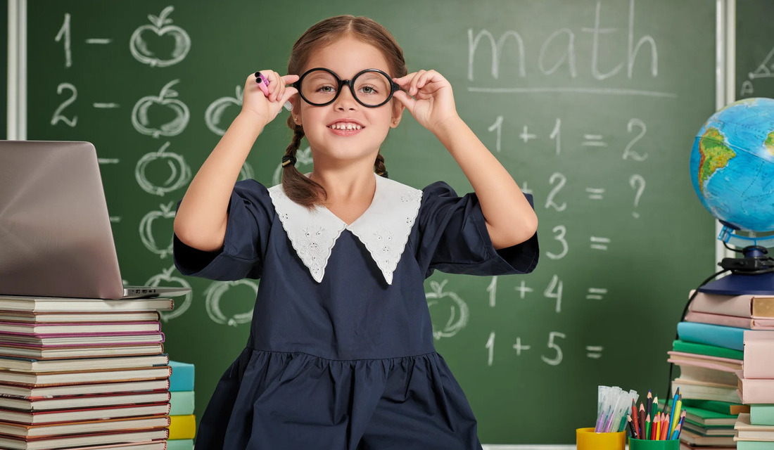 A young girl wearing glasses sits at a desk in a classroom, focused on her work.