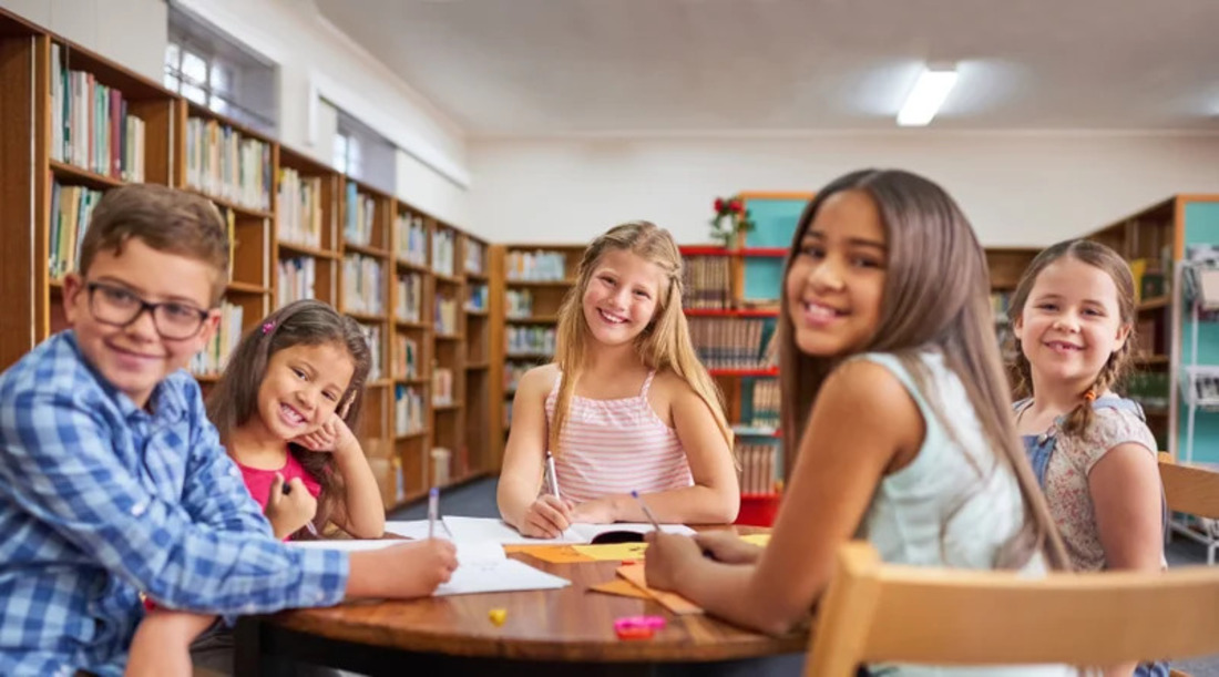 A group of children seated at a library table, focused on their books and sharing ideas.