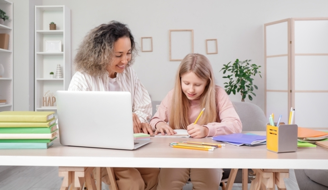 A woman and a girl sit together at a table, focused on a laptop screen in front of them.