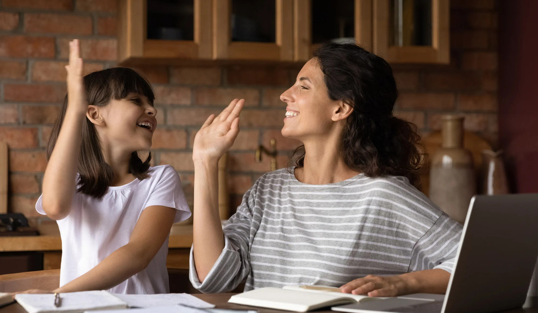A woman and a child sitting at a table, both raising their hands in a joyful gesture.