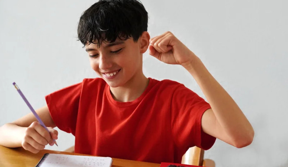 A young boy at a desk raises his arms, likely celebrating or seeking attention.
