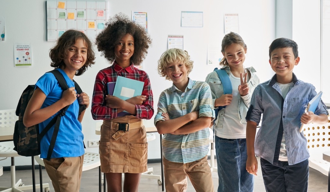 A group of children stands in a classroom, each wearing backpacks and engaged in conversation.