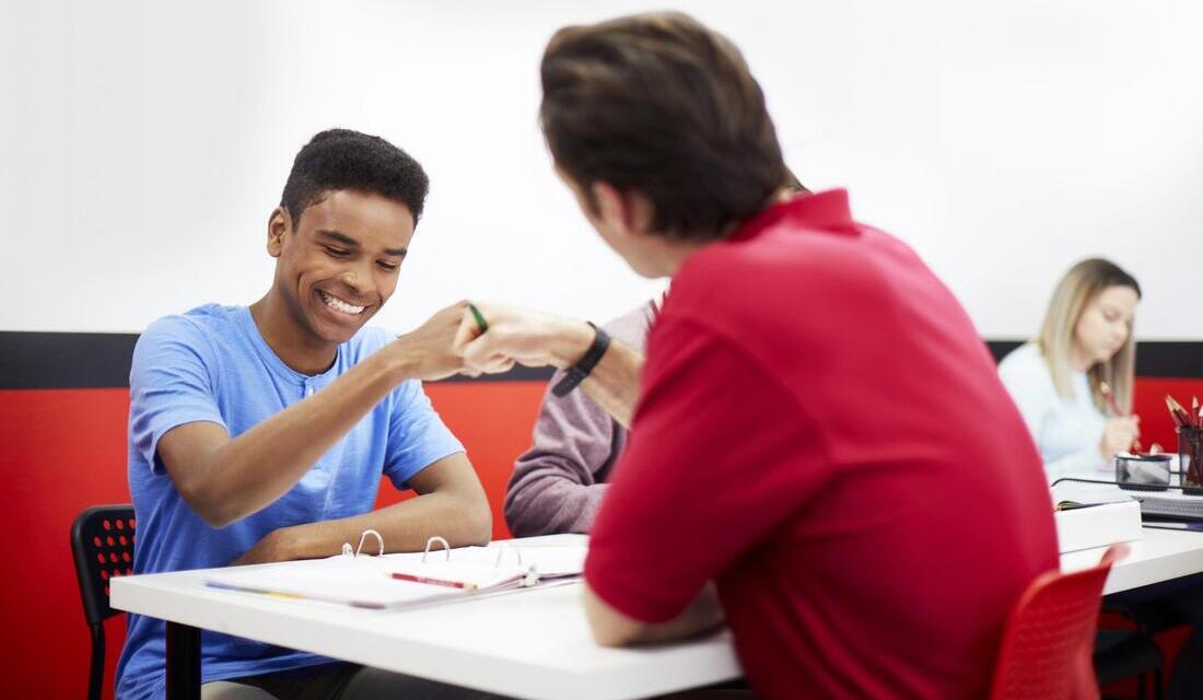A teacher and a student collaborate at a table, both focused on a pen and materials in front of them.