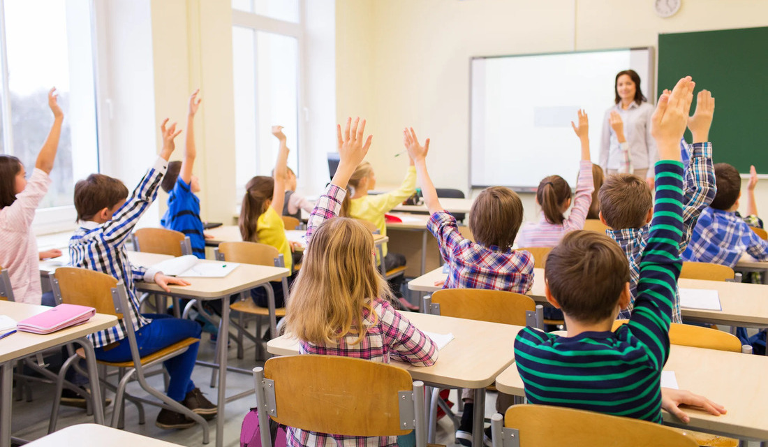 Several children in a classroom enthusiastically raising their hands, indicating their desire to contribute to the lesson.