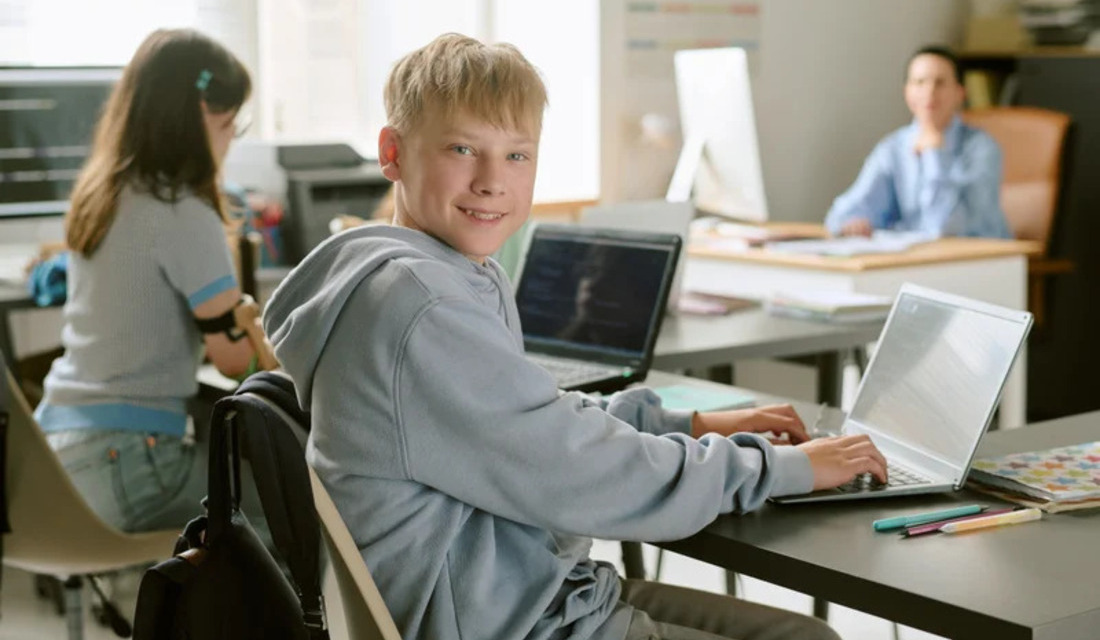 A young boy seated at a desk, focused on his laptop, with books and stationery nearby.