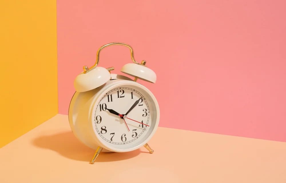 A vintage-style analog alarm clock on a colorful orange and pink background, showing approximately 10:08.
