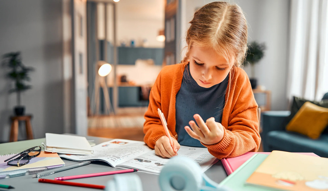 A young girl sits at a table, focused on writing in her notebook with a pencil in hand.