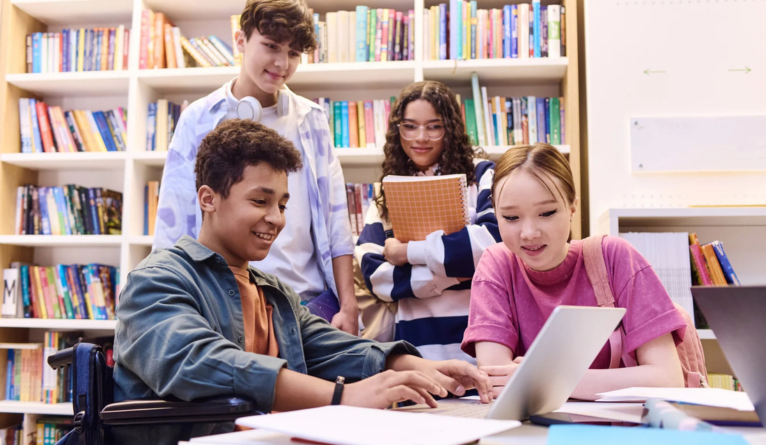 A group of students studying together in a library, focused on a laptop and surrounded by books.