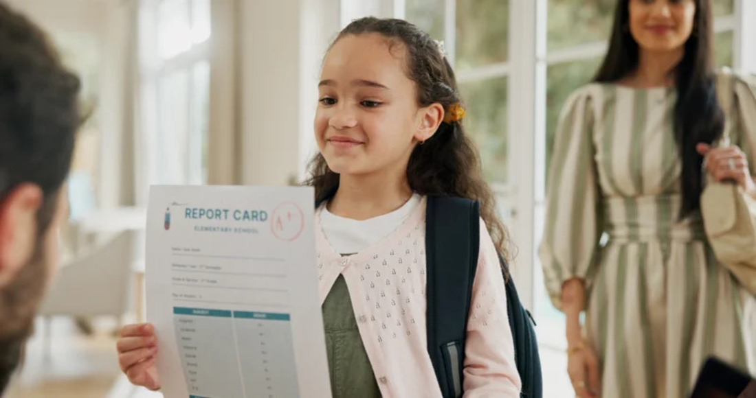 A young girl shows her school report, smiling as she presents her academic performance on a piece of paper.
