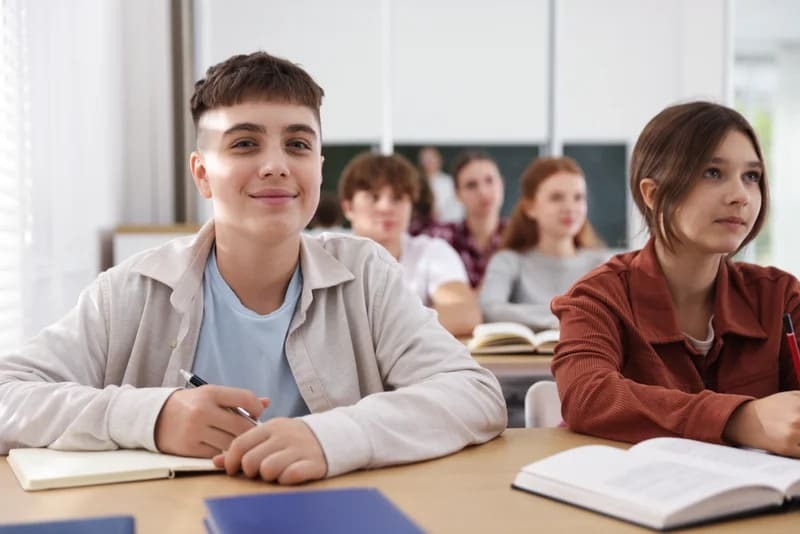 A group of teenage students engaged in a classroom discussion, seated at desks with books and laptops.