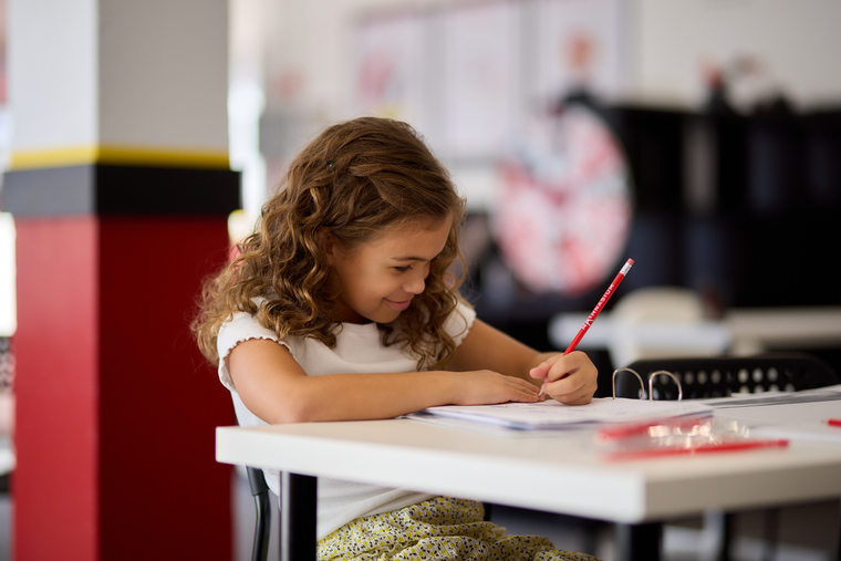 A young girl sits at a table, focused on writing in a notebook with a pencil in hand.