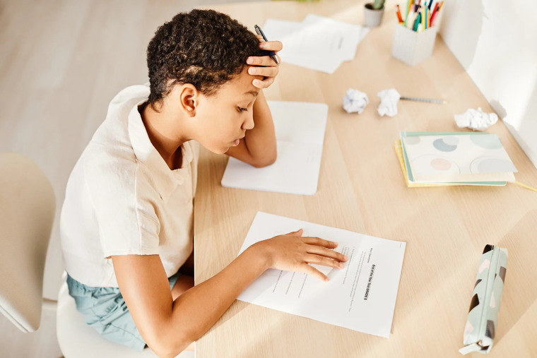 A child rests his head on his hand while sitting at a desk, appearing thoughtful or tired.