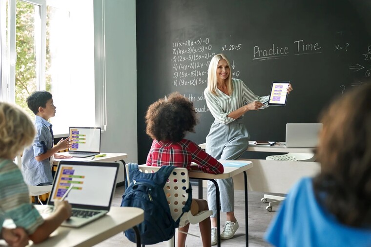 A teacher instructs a diverse group of students seated at desks in a bright classroom setting.