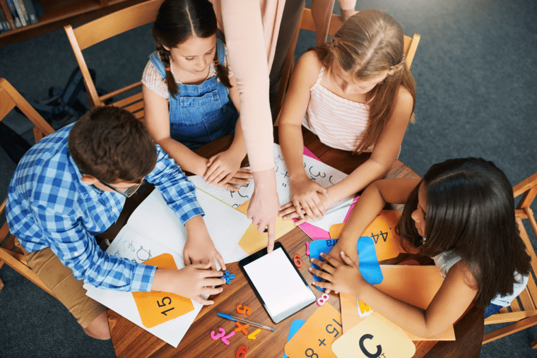 A group of children collaborating at a table with a tablet and sheets of paper.
