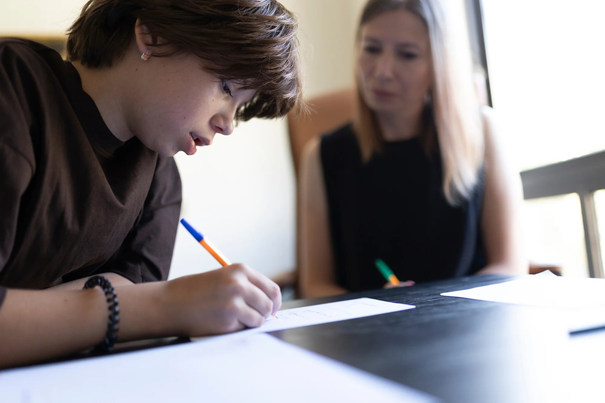 A young boy writes on paper while a woman observes him attentively.
