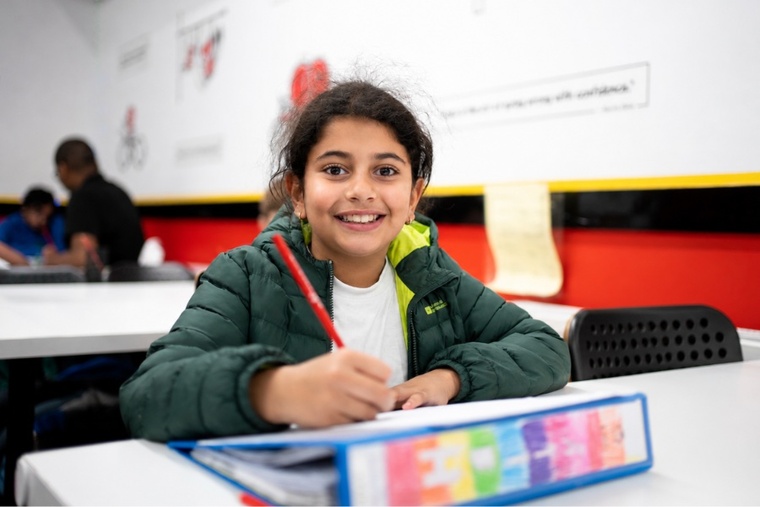 A girl smiles while sitting at a table, holding a pen and paper, engaged in writing or drawing.