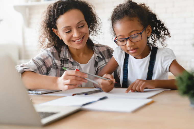 A mother and daughter sit at a table, collaborating on homework with books and papers spread out before them.