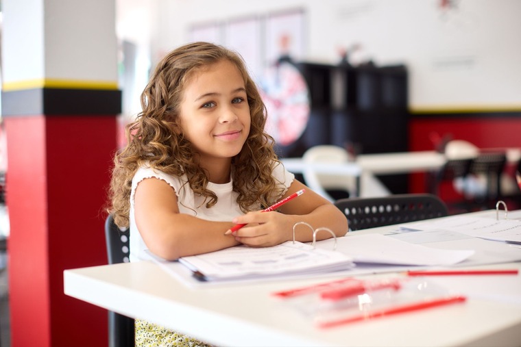 A young girl sits at a table, focused on writing with a pen on a sheet of paper.