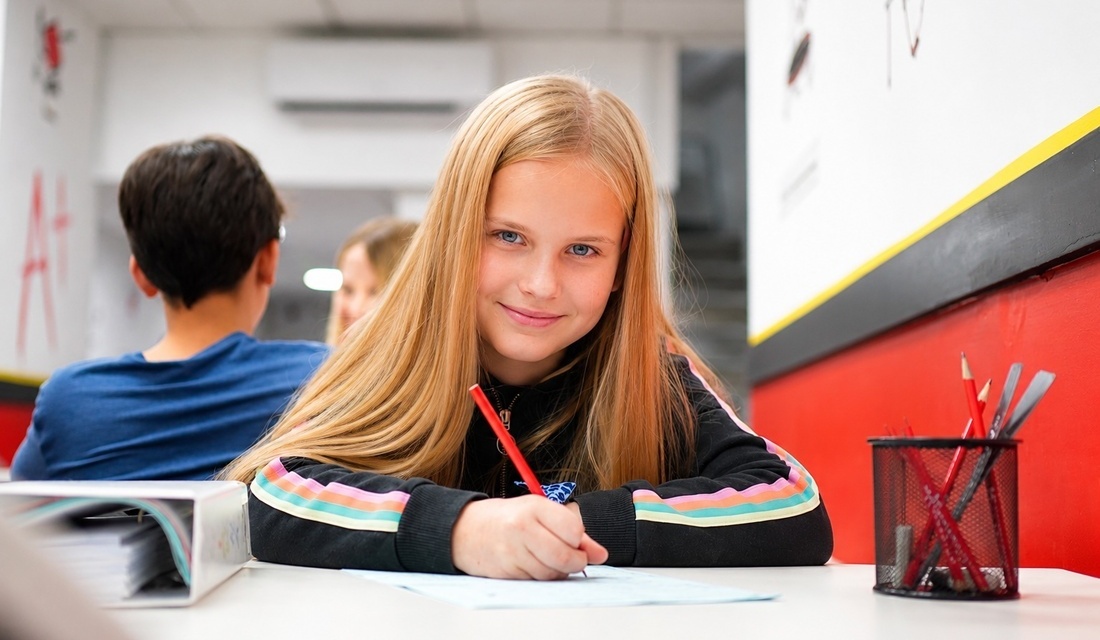 A girl sits at a table, focused on writing in a notebook with a pen in her hand.