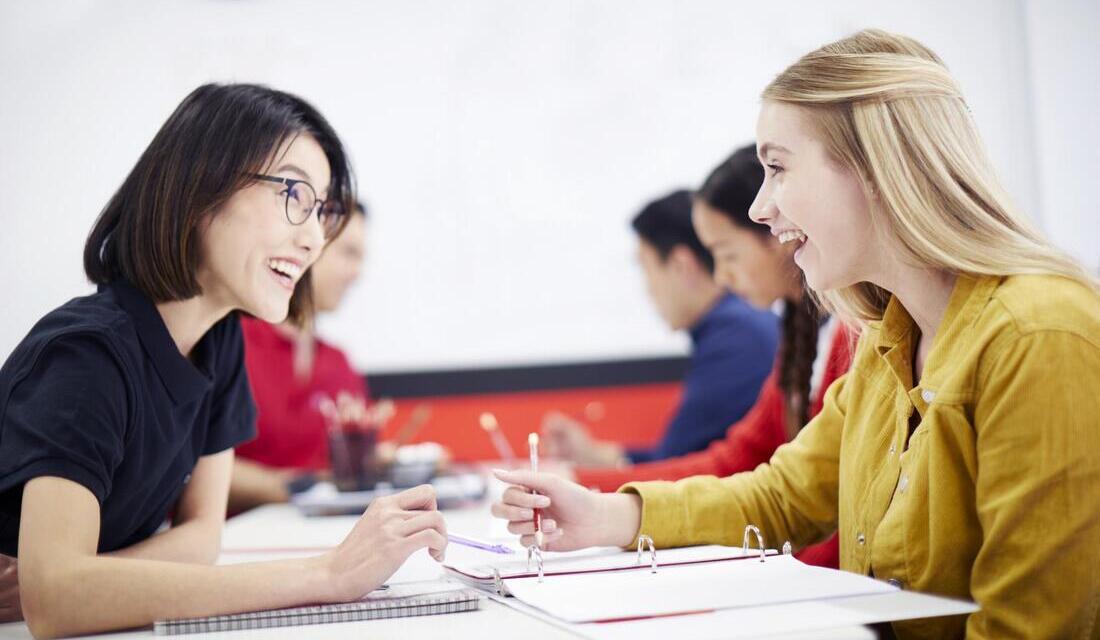 A math tutor and a student engage in discussion at a classroom desk, surrounded by books and learning materials.