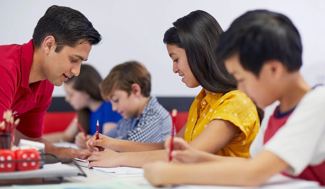 A man and woman instruct a group of children in a bright classroom filled with educational materials and colorful decorations.