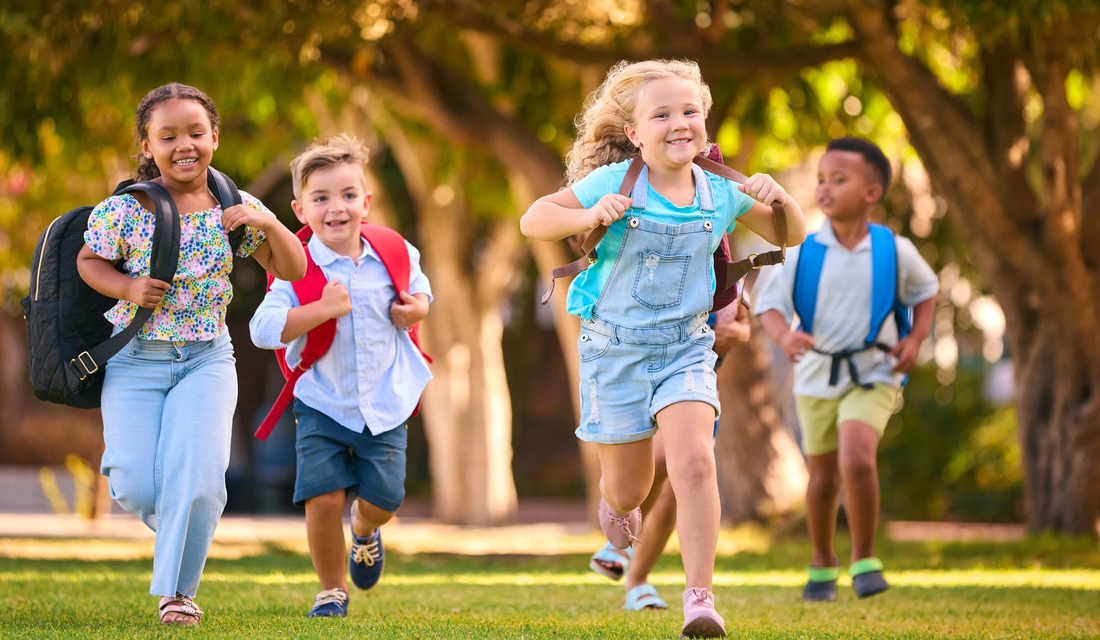 Children joyfully running in a park, each carrying colorful backpacks, surrounded by green grass and trees.